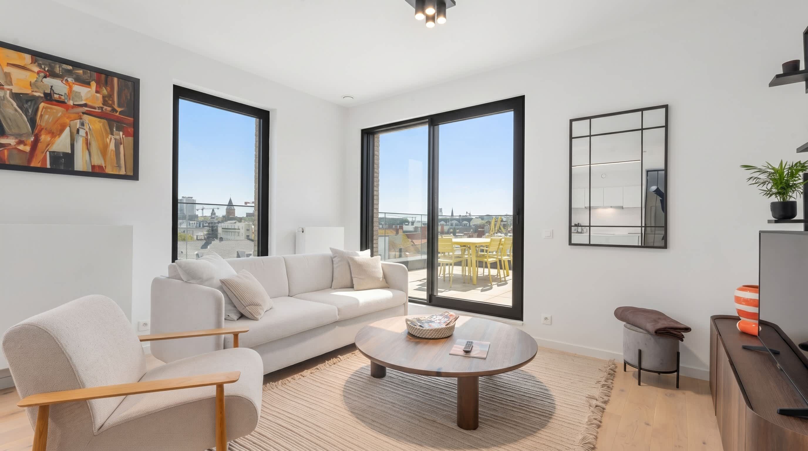 Ein modernes Wohnzimmer mit Sofa, Sessel, Couchtisch und großen Fenstern mit Blick auf die Stadt.
