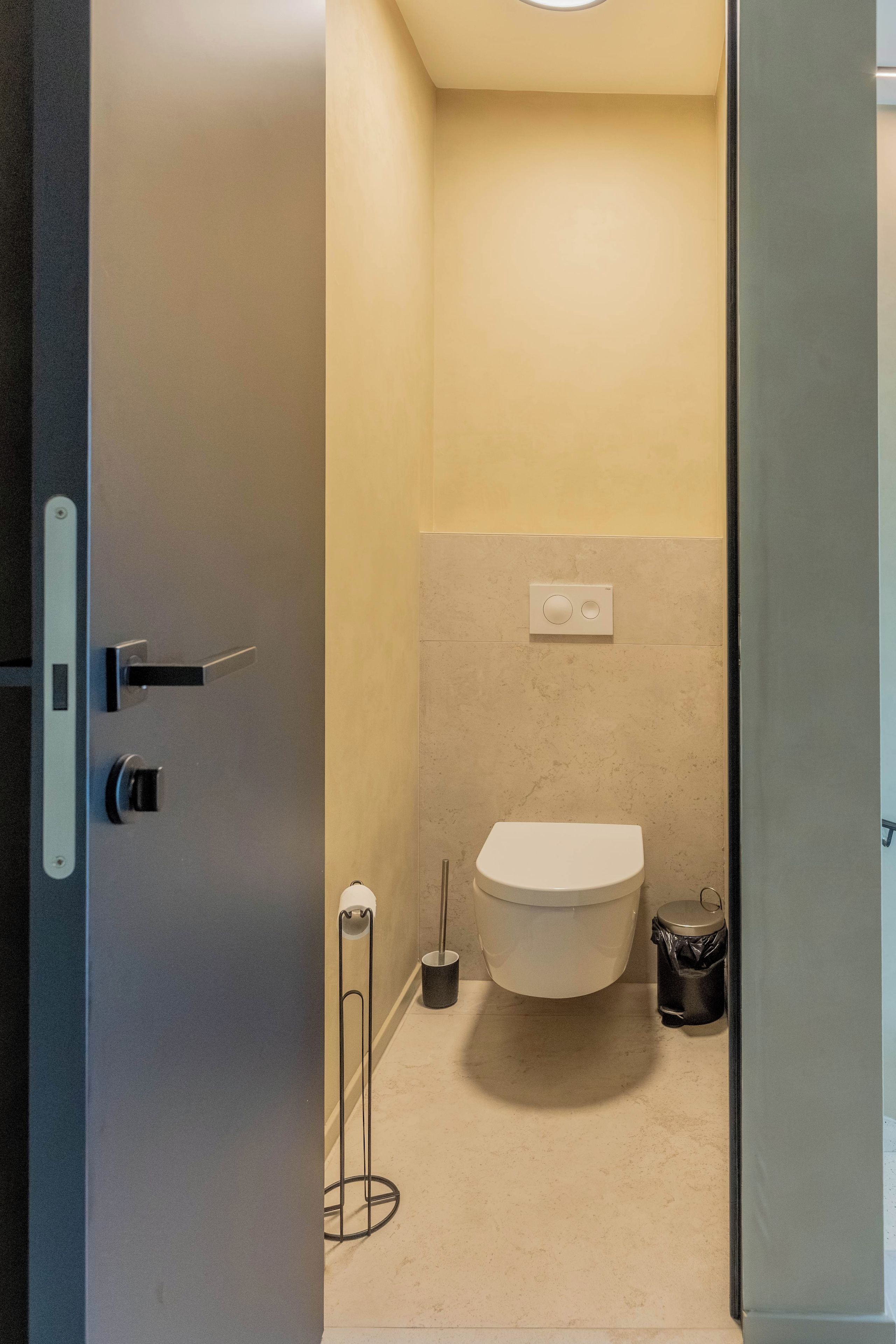 Un baño pequeño y moderno con inodoro, un accionador de descarga en la pared y un cubo de basura.