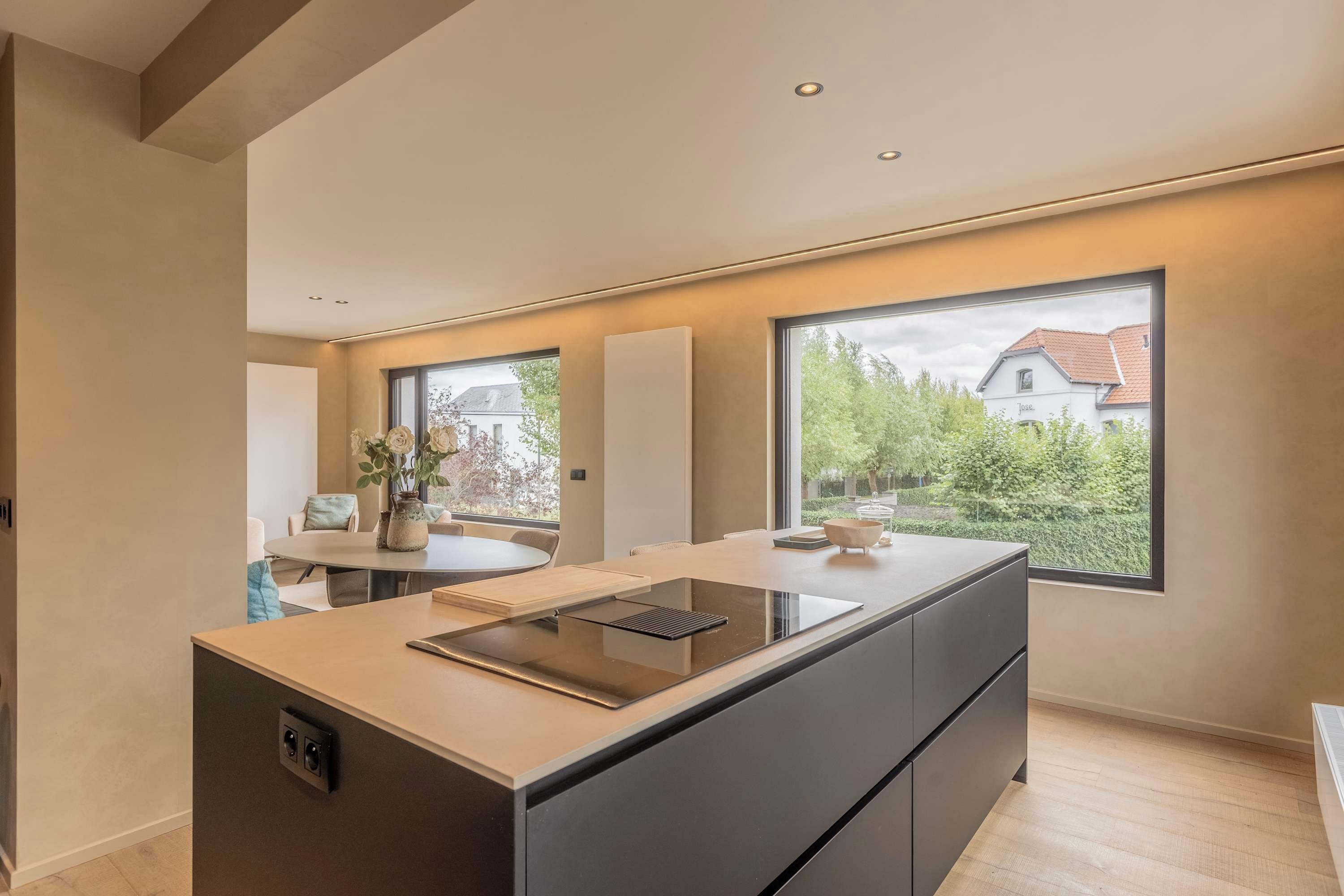 Eine moderne Küche mit einer zentralen Kochinsel, großen Fenstern und Blick auf ein Haus im Freien.