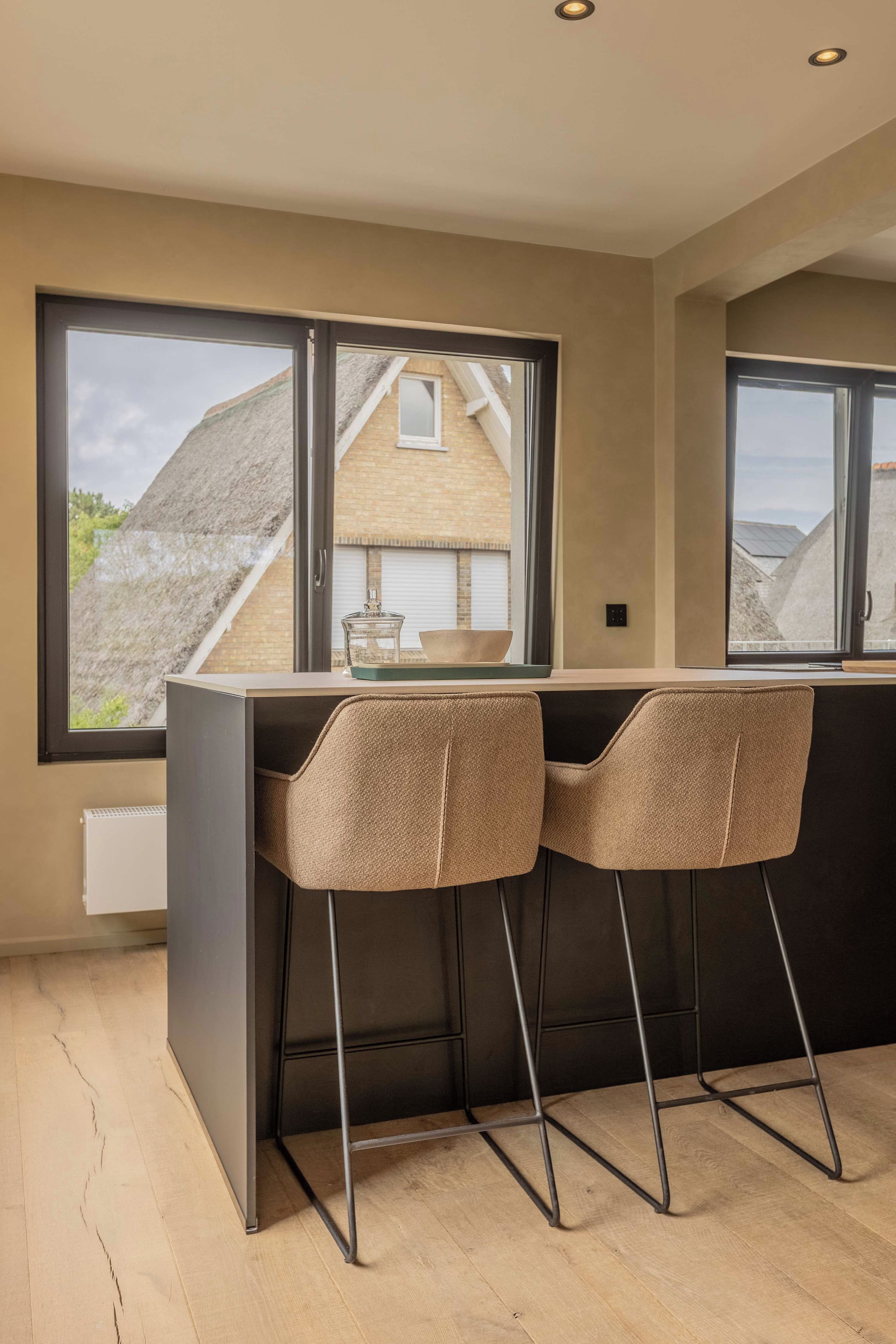 Eine moderne Küche mit Barbereich, zwei hohen Barhocker und großen Fenstern mit Blick auf eine Wohngegend.