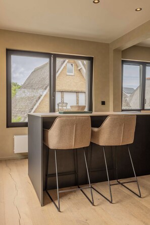 A modern kitchen with a bar area, two high stools, and large windows overlooking a residential area. - Villa Hélène · bright upstairs flat, 3 bedrooms (Koksijde)