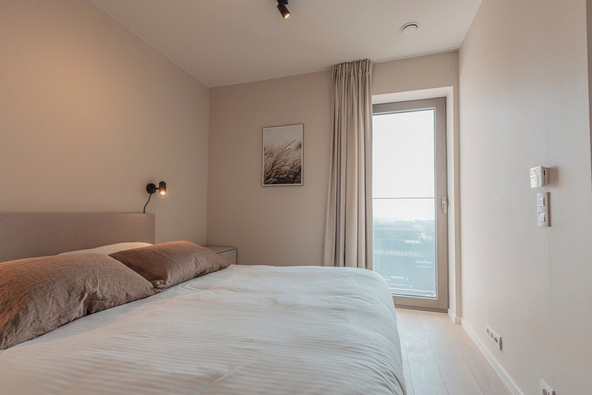 A modern bedroom with a large bed, a headboard, a bedside lamp, a window with curtains, and a wall-mounted artwork.