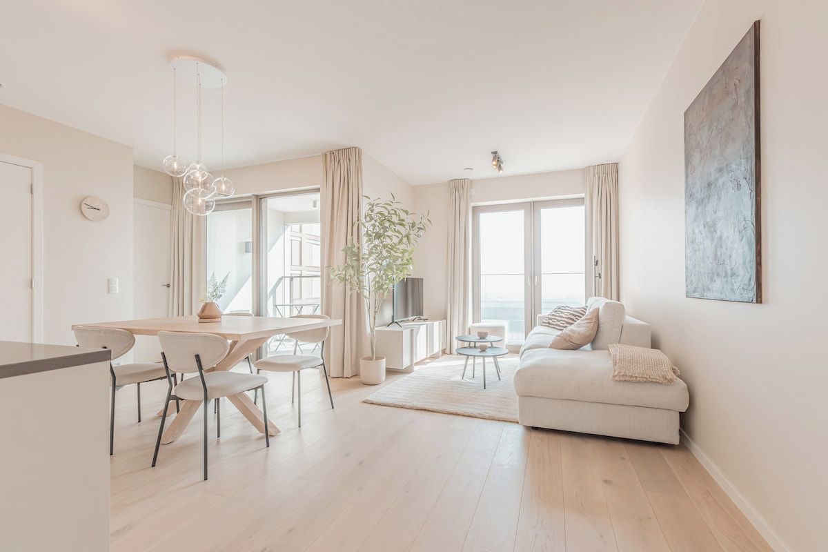 A modern living room with a sofa, a dining table with chairs, a TV, and a large window.