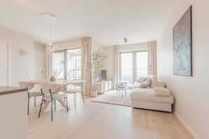 A modern living room with a sofa, a dining table with chairs, a TV, and a large window. - Seven O'Sea · 7th-floor flat, beach 3 min away (Ostend)