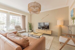 Moderni olohuone, jossa on nahkasohva, televisio, puinen sohvapöytä ja suuri ikkuna, jossa on verhot.