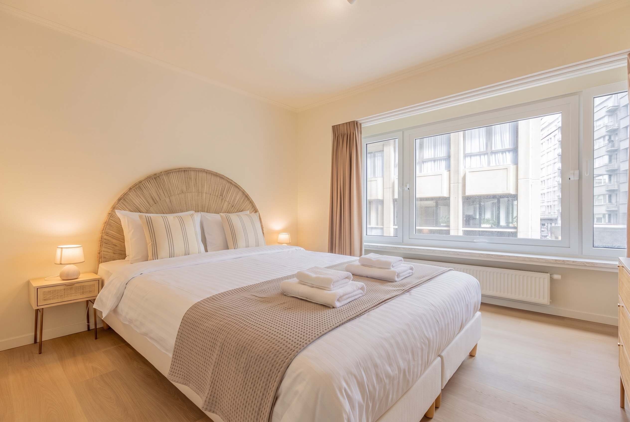 Hotellihuone, jossa on suuri sänky, yöpöytä, ikkuna verhoilla ja näkymä ulkona oleviin rakennuksiin.