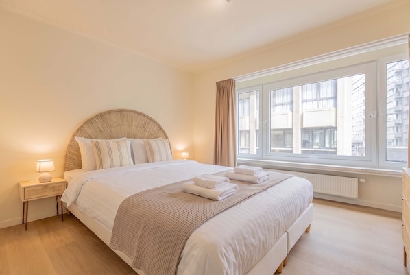 Hotellihuone, jossa on suuri sänky, yöpöytä, ikkuna verhoilla ja näkymä ulkona oleviin rakennuksiin.