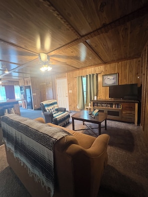 A cozy living room with wooden walls, a sofa, a coffee table, and a television. - Cozy 2-bedroom mobile home in adorable Chouteau with AC (Chouteau)