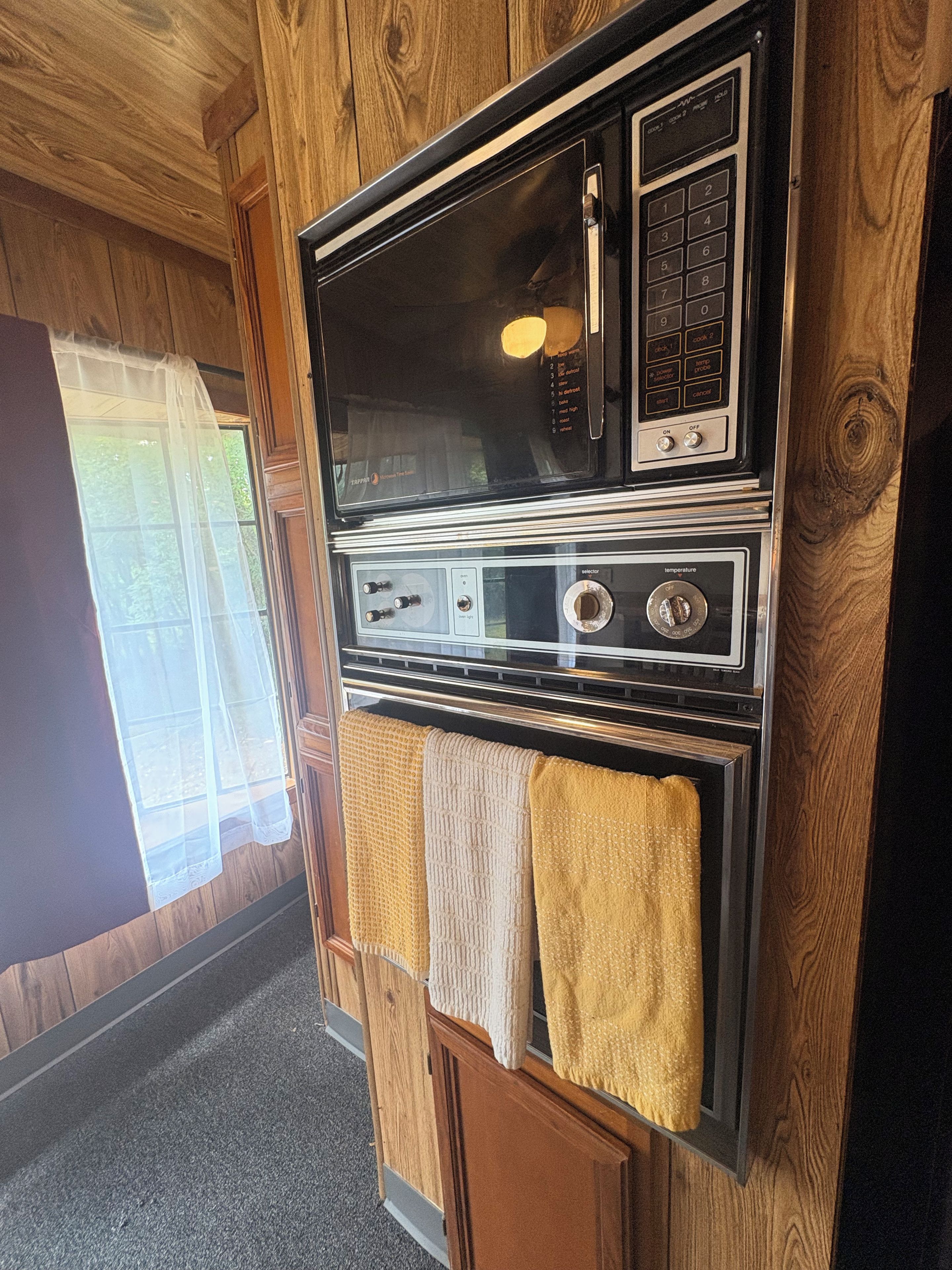 Una cocina con horno integrado y microondas, paneles de madera y un ventanal con cortinas traslúcidas.