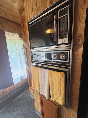 Una cocina con horno integrado y microondas, paneles de madera y un ventanal con cortinas traslúcidas.