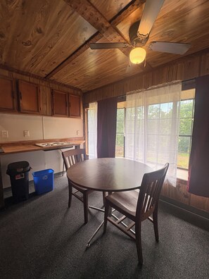 A wooden interior with a round table and chairs, a ceiling fan, and a window with curtains. - Cozy 2-bedroom mobile home in adorable Chouteau with AC (Chouteau)