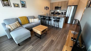 A modern living room with a grey sofa, a wooden coffee table, and a kitchen area with a stainless steel refrigerator and microwave.