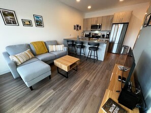 Un salon moderne avec un canapé gris, une table basse en bois et un coin cuisine doté d’un réfrigérateur et d’un four à micro-ondes en acier inoxydable.