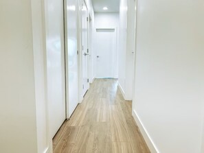 Un couloir aux murs blancs et au plancher en bois, qui mène à une porte au fond.