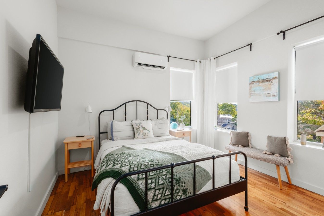 A bedroom with a bed, bedside tables, a TV, an air conditioning unit, and a window with a view.