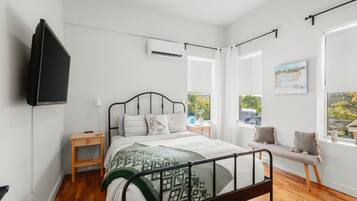 A bedroom with a bed, bedside tables, a TV, an air conditioning unit, and a window with a view.