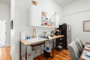 Cuina compacta amb un rentaplat, una placa de cocció i un frigorífic. Hi ha una taula de dinar amb cadires i una petita prestatgeria sobre el rentaplat.