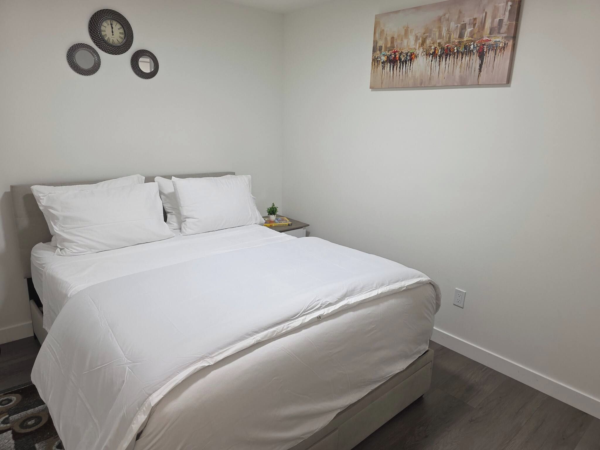 Una cama bien hecha con sábanas blancas en una habitación con pared lisa y un cuadro.
