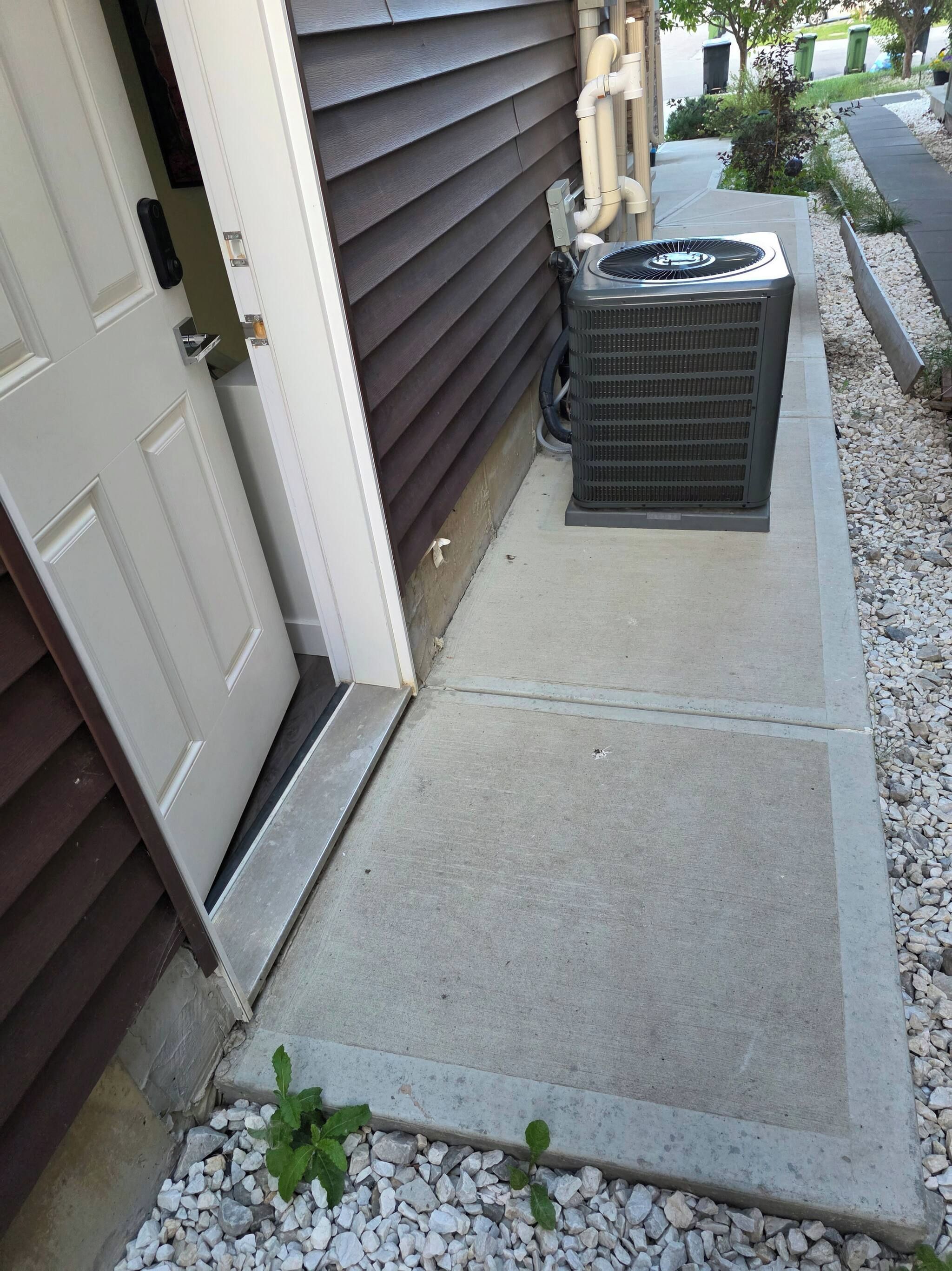 Un vestíbulo residencial con un escalón de concreto que lleva a una puerta, una unidad de aire acondicionado pequeña y un sendero de grava.