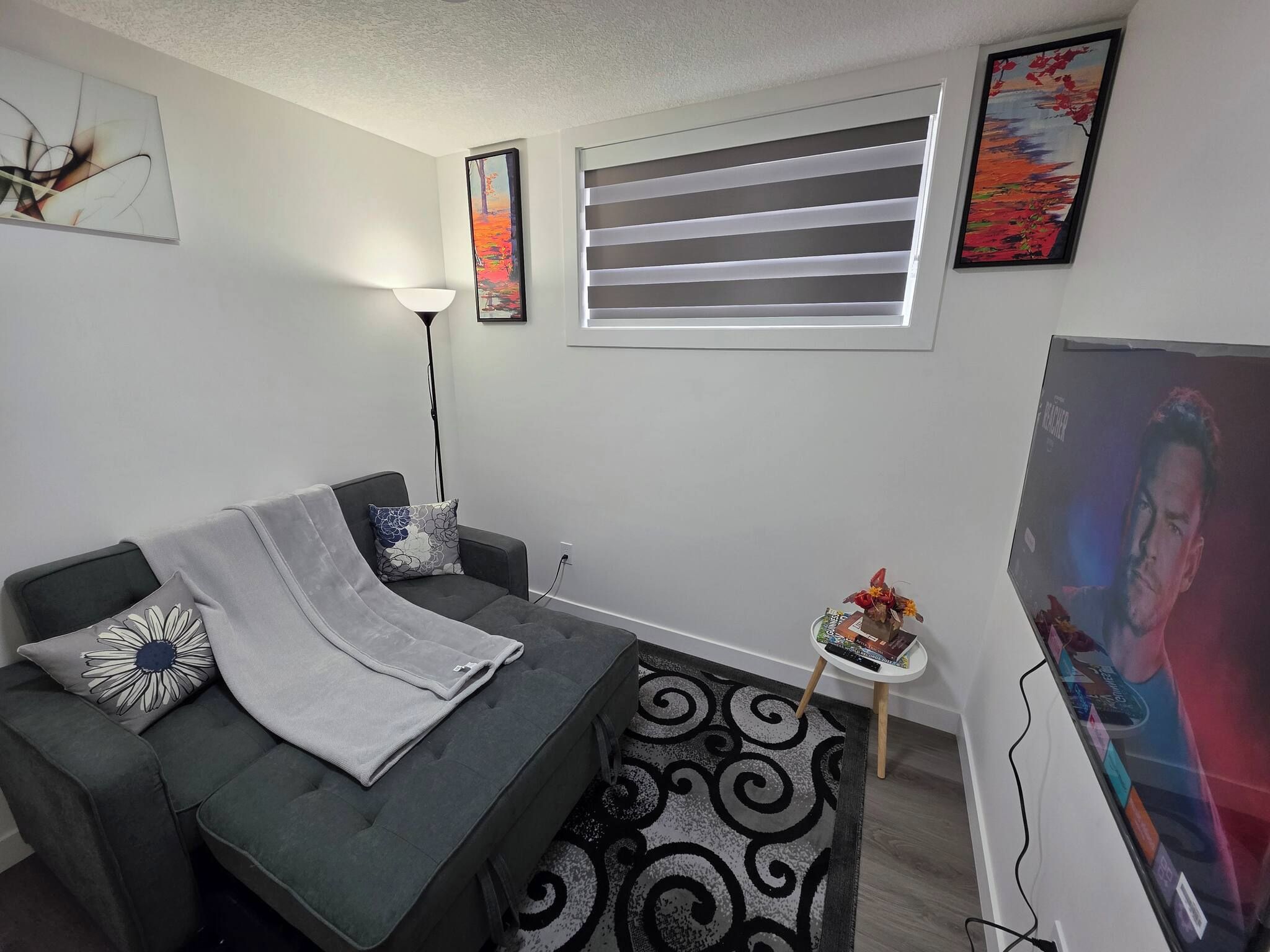 A living room with a grey sofa, a floor lamp, a window with blinds, and a TV on a stand.