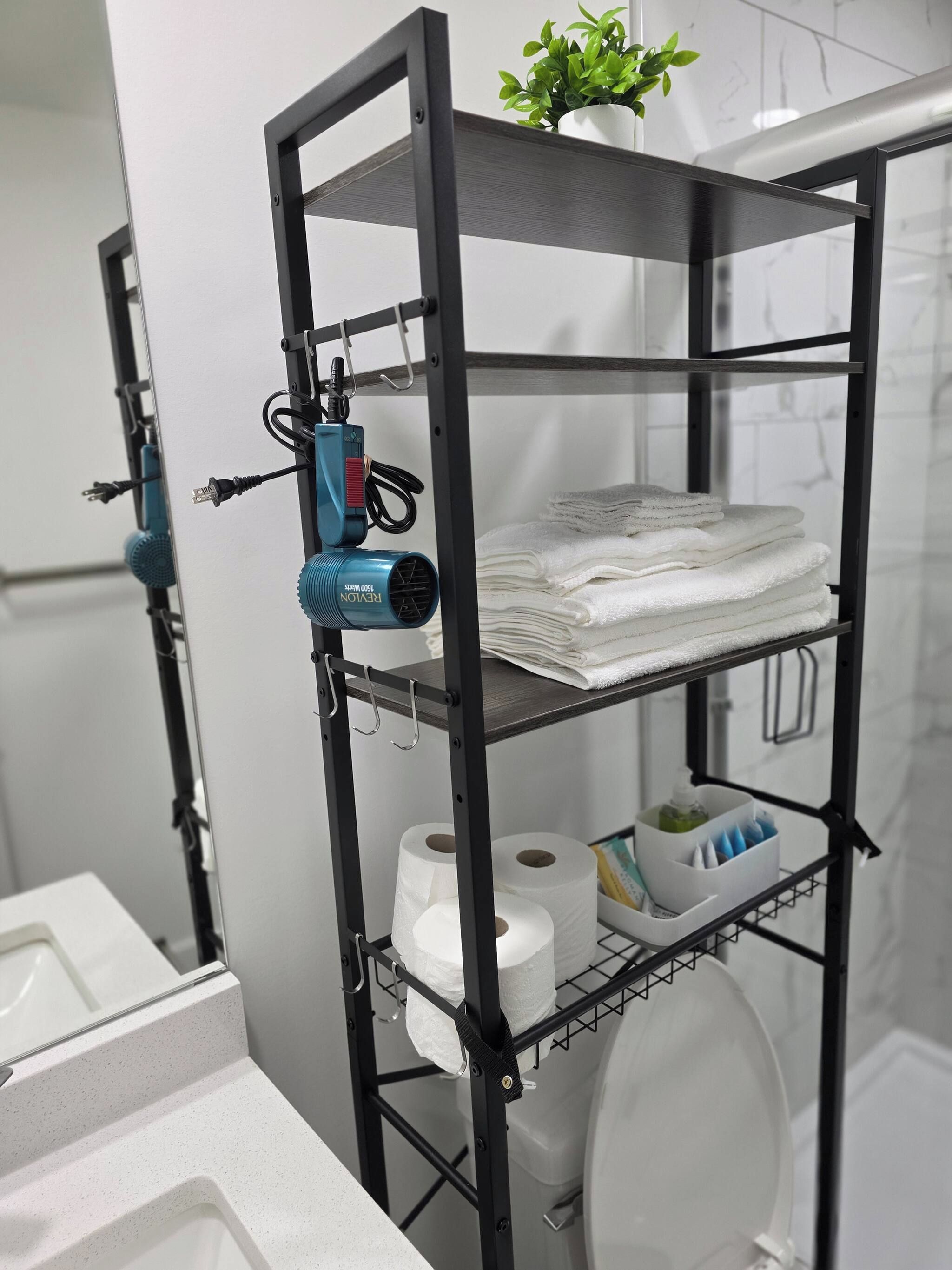 Un baño con un estante metálico negro que contiene toallas, papel higiénico y un secador azul.