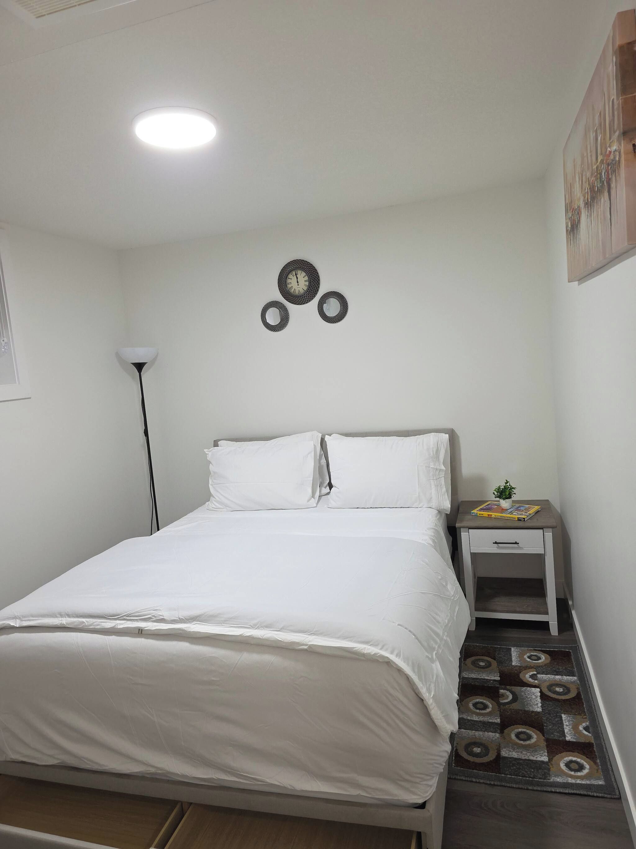 Una cama bien tendida con ropa de cama blanca, una mesita de noche con una planta y libros, y un reloj de pared.