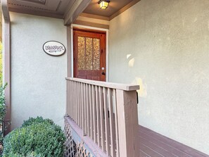 Una porta in legno con un pannello di vetro decorativo, un piccolo portico con ringhiera in legno e un cartello sopra la porta.
