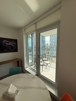 Een kamer met een eettafel, stoelen, een schilderij aan de muur en uitzicht op hoge gebouwen door grote glazen deuren.