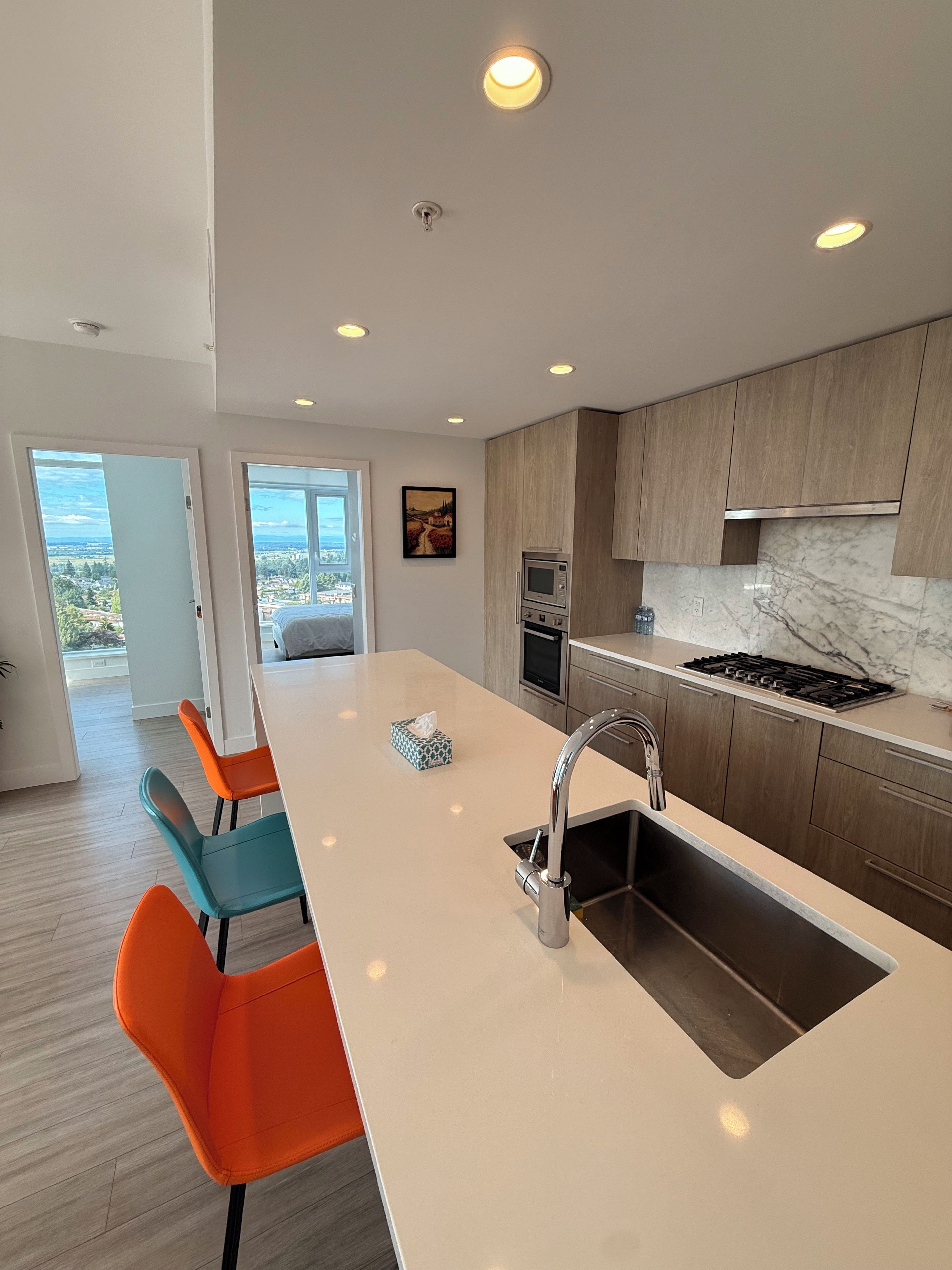 A modern kitchen with a central island, stainless steel appliances, and a view through an open door.