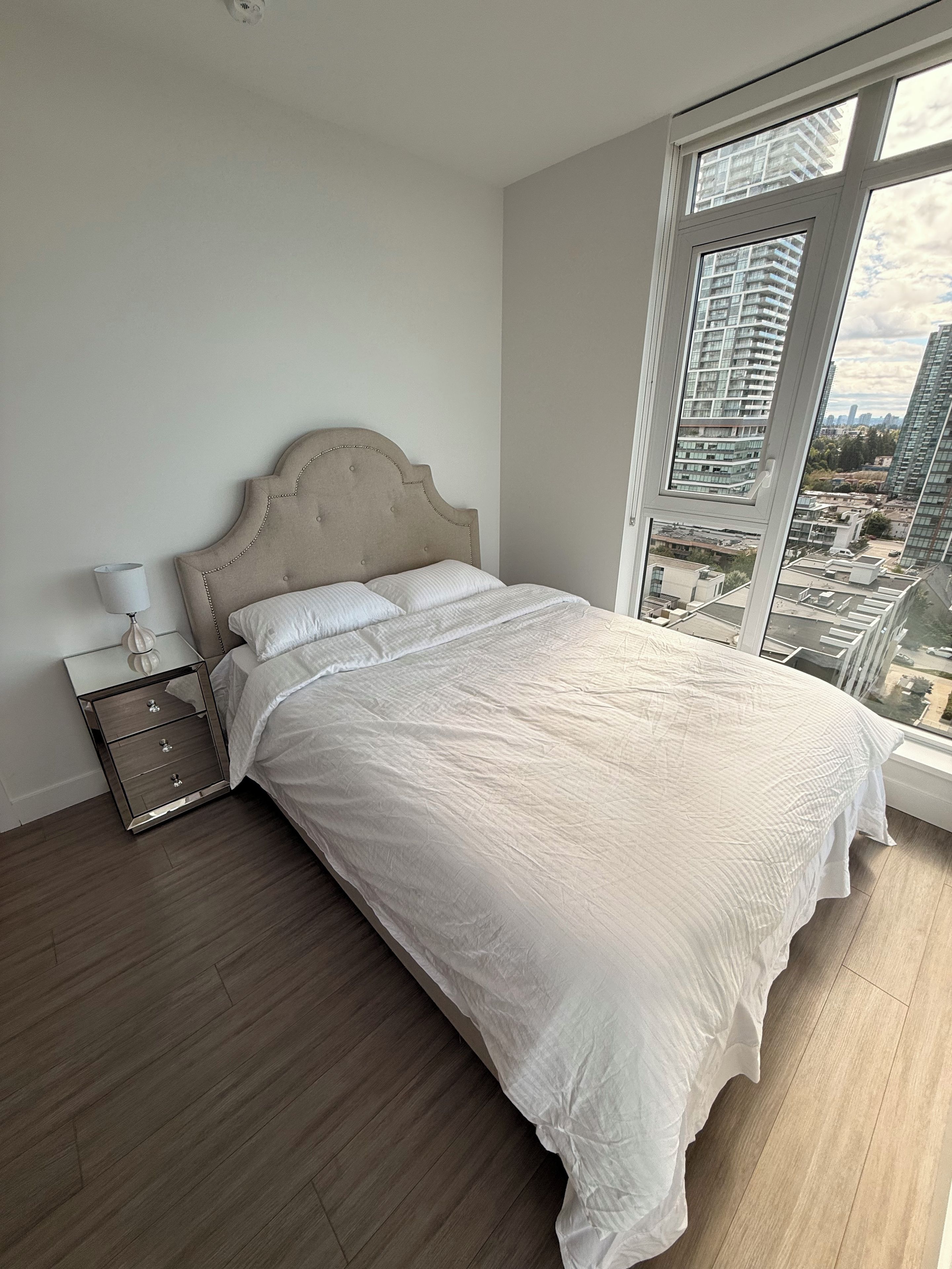 A bedroom with a large bed, a nightstand, a lamp, and a window with a city view.