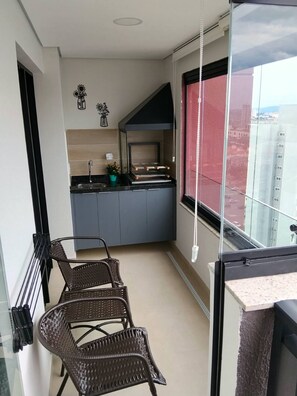 Un petit balcon agrémenté de chaises en osier, une petite cuisine avec des meubles gris et une vue sur la ville par la fenêtre.