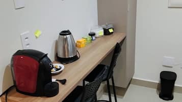 Une petite cuisine équipée d’un plan de travail en bois, d’une bouilloire, d’une machine à café et de quelques récipients. On y trouve également deux chaises noires et une poubelle noire.