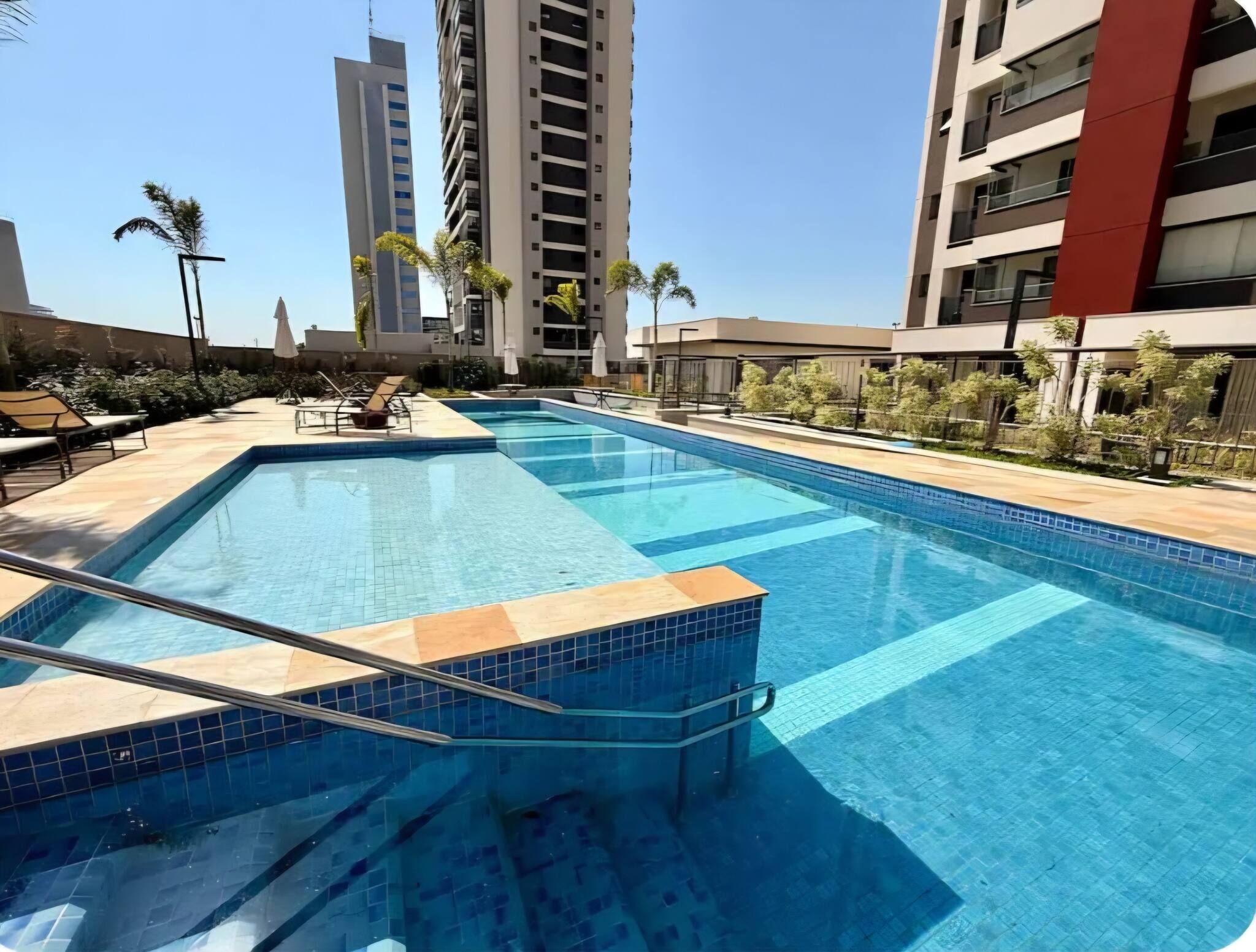 Una piscina rectangular con borde de azulejos, rodeada de sillas de descanso y sombrillas, con edificios altos al fondo.