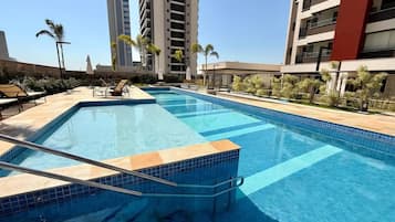Una piscina rectangular con borde de azulejos, rodeada de sillas de descanso y sombrillas, con edificios altos al fondo.