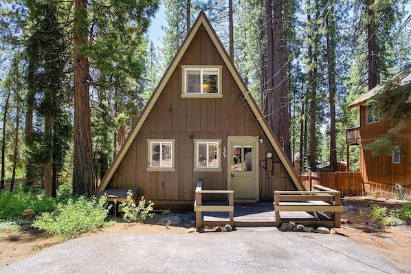A wooden cabin with a triangular roof, two windows, and a door, surrounded by a forest.