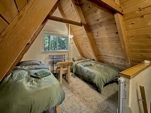 A small, cozy attic bedroom with two beds, a desk, and a window overlooking a forest.