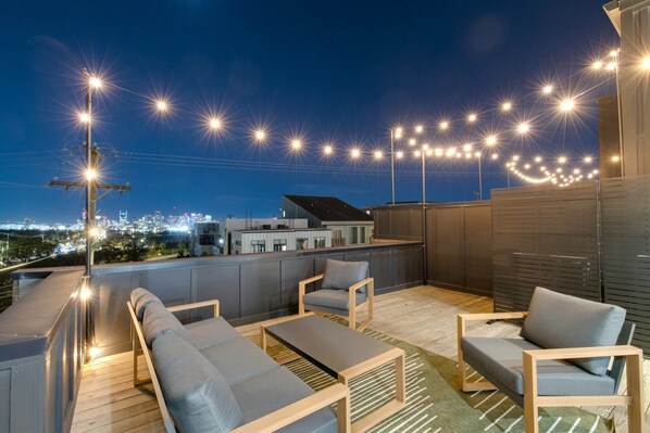 An outdoor seating area with sofas and a table, string lights overhead, and a cityscape view in the background.