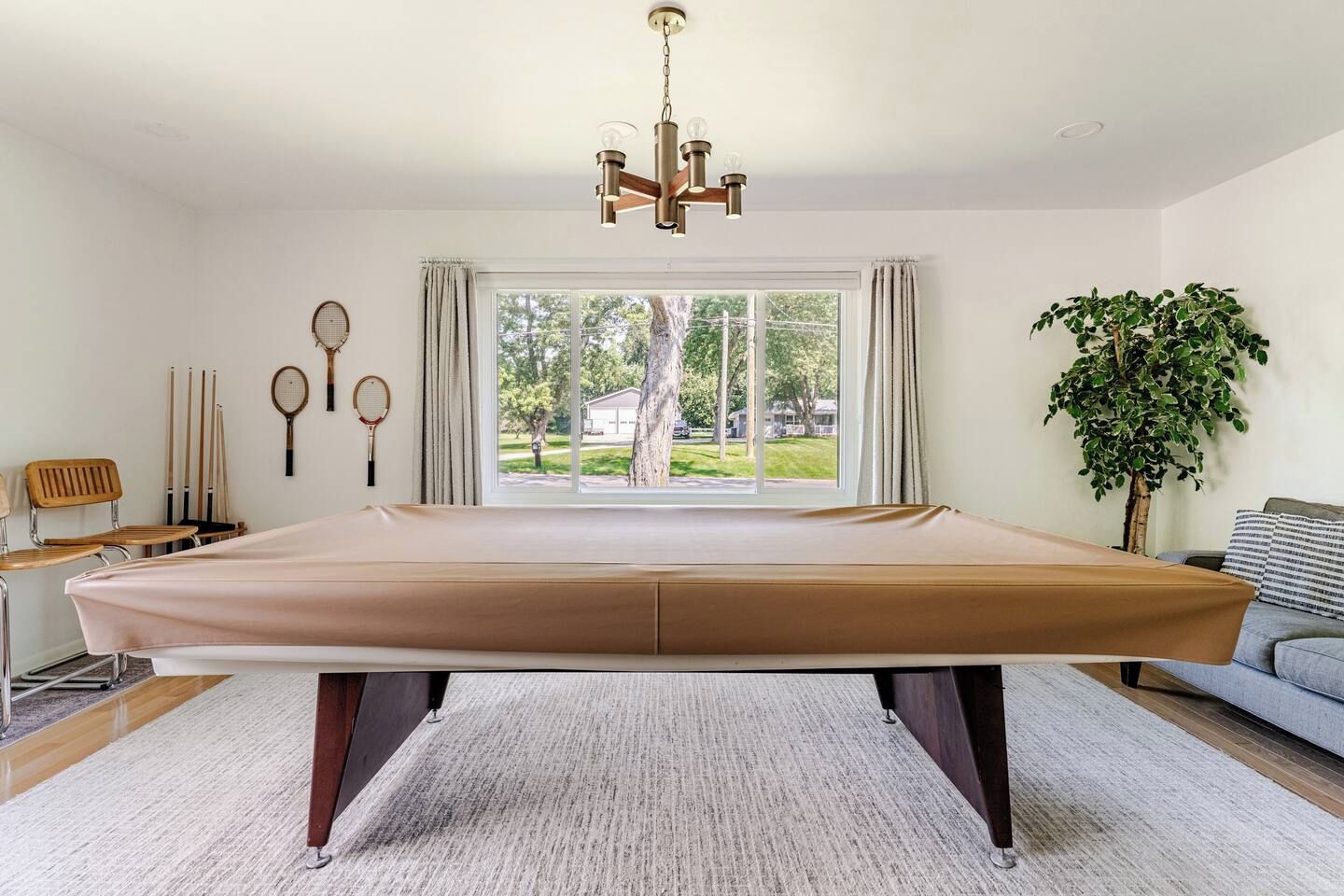 A spacious room with a pool table, a large window with curtains, a hanging light fixture, and a potted plant.