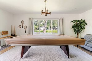 A spacious room with a pool table, a large window with curtains, a hanging light fixture, and a potted plant. - Atomic Acres- Pool, Hot tub, Fire pit & more (Sawyer)