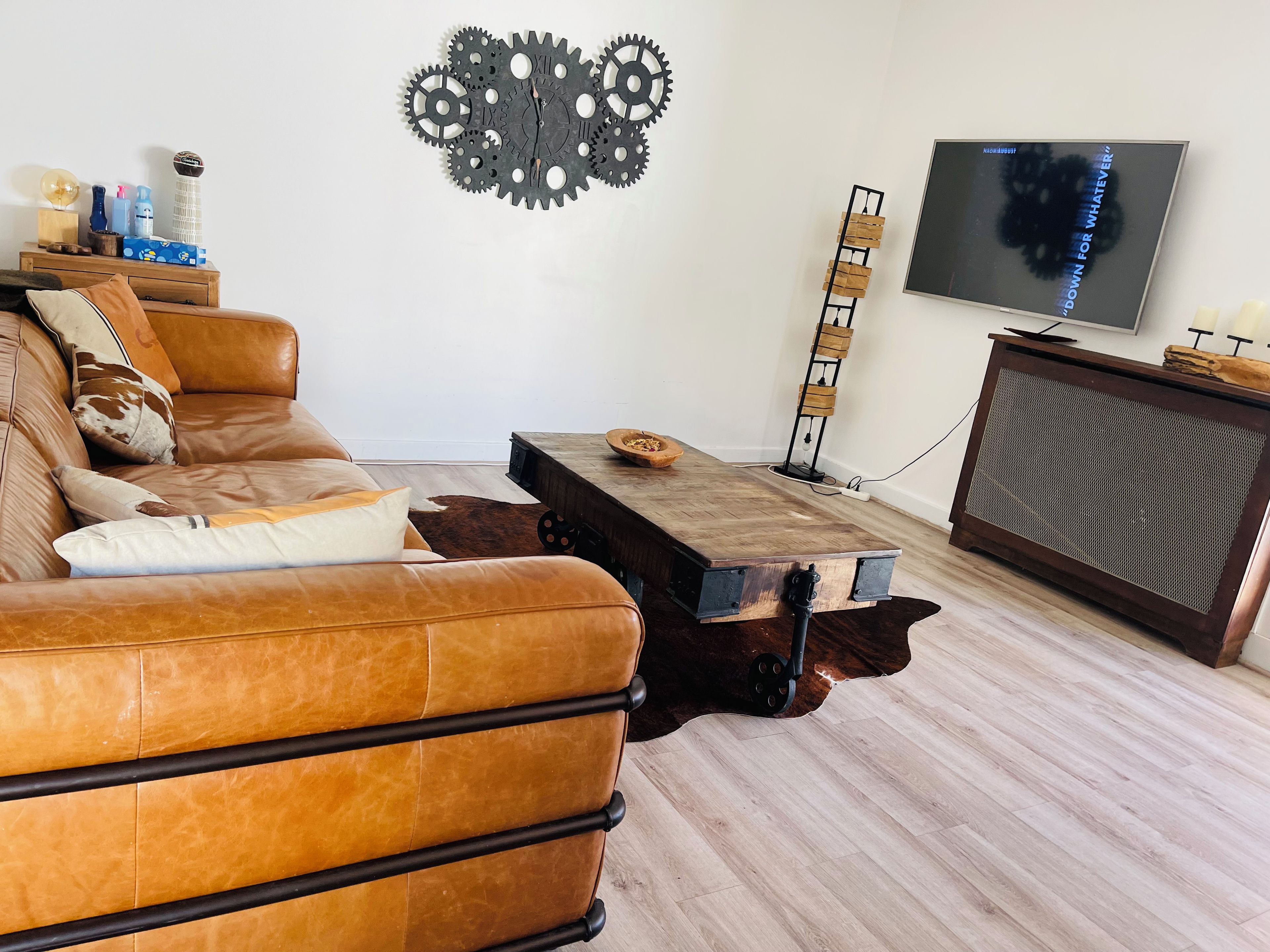 Un salon avec un canapé en cuir marron, une table basse en bois, un téléviseur fixé au mur et un élément décoratif en forme d’engrenage accroché au mur.