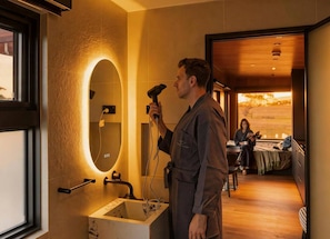 Une personne utilise un sèche-cheveux dans une pièce dotée d’un grand miroir, d’un plancher en bois et d’une fenêtre offrant une vue sur le coucher du soleil.