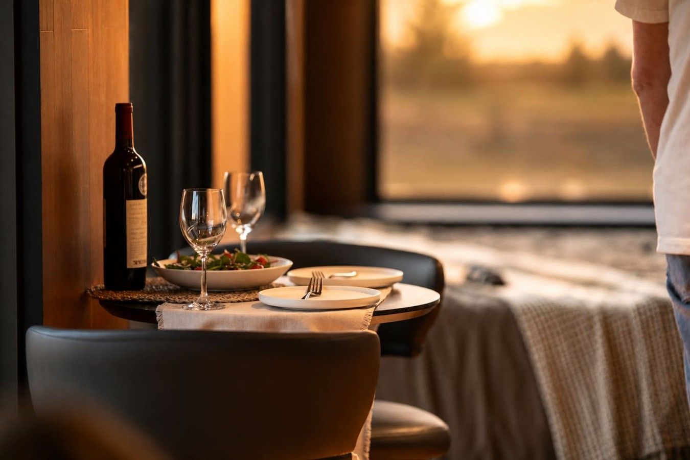A dining table set with a bottle of wine, glasses, and plates, with a view of a sunset through a window.