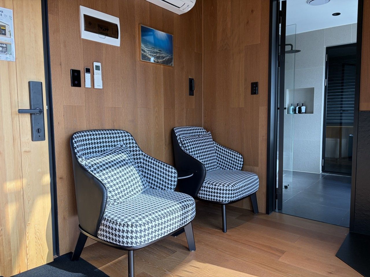 Deux fauteuils à motif pied-de-poule dans un salon moderne avec des lambris en bois et une porte en verre donnant sur un espace douche.