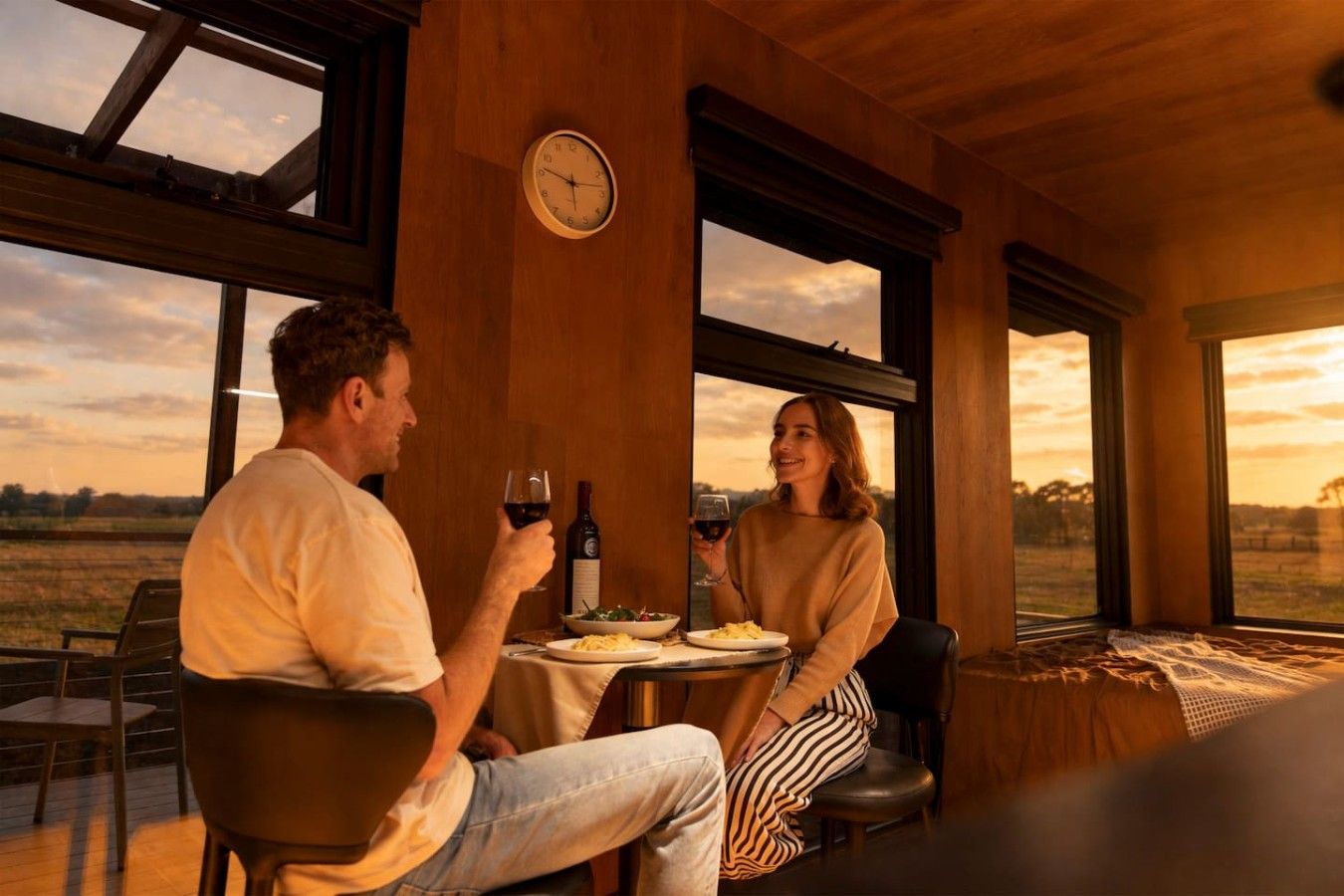 Et par spiser ved et bord med udsigt over landskabet, et ur på væggen og en flaske vin.