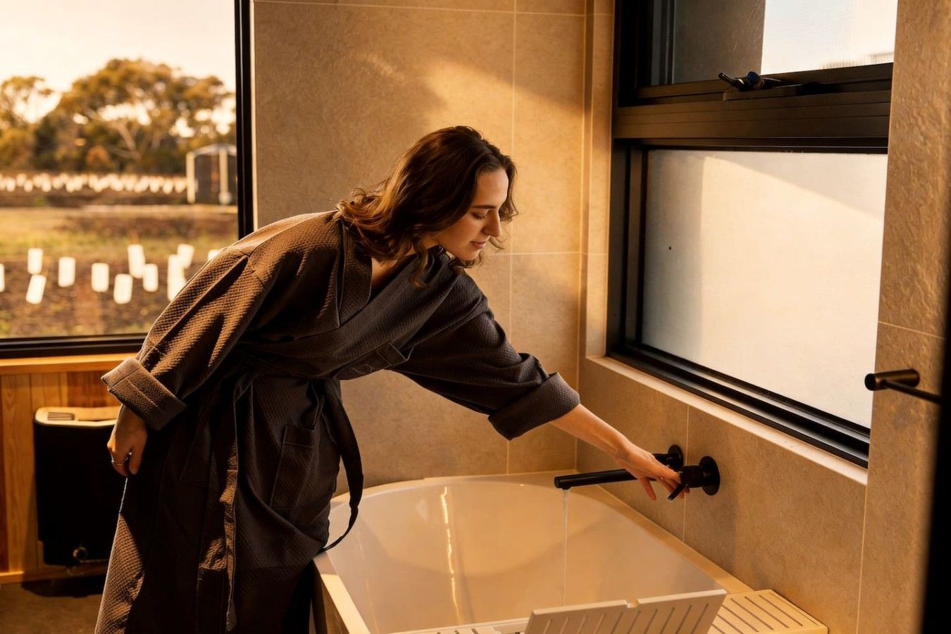 A person in a bathrobe reaching for a bathtub faucet.