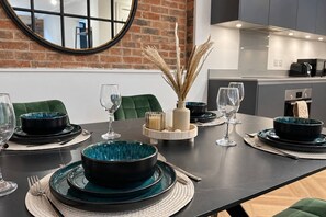 Een eettafel gedekt met zwart en teal servies, een vaas met gedroogde planten en een moderne keuken op de achtergrond.