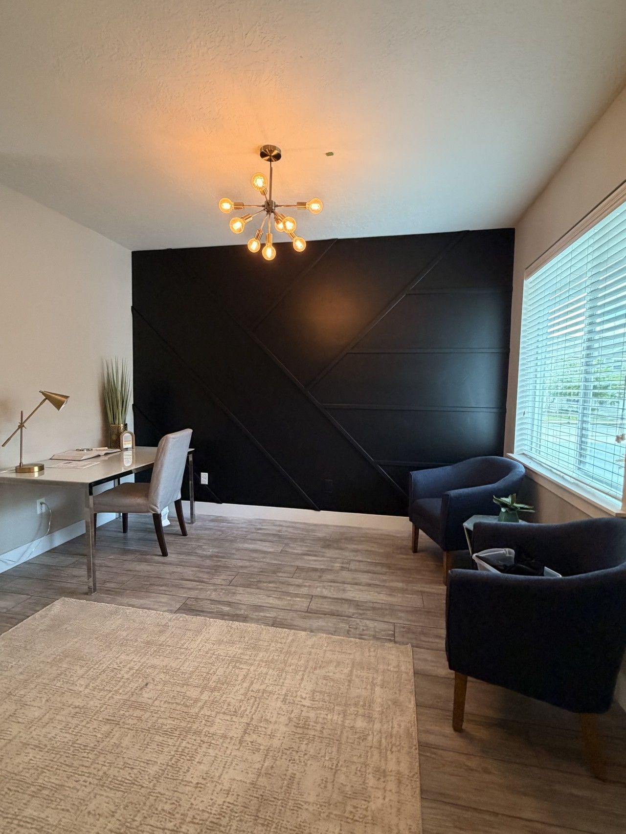 A modern living room with a dark accent wall, a desk with a chair, two armchairs, a floor lamp, and a large window with blinds.