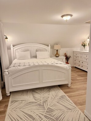 Un dormitorio con una cama blanca, una mesita de noche, un tocador y una lámpara.