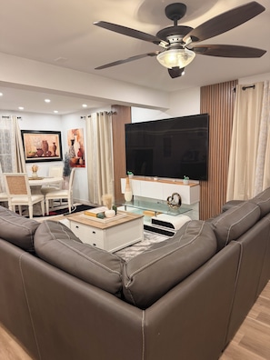 A modern living room with a brown leather sofa, a glass coffee table, a flat-screen TV, and a ceiling fan.