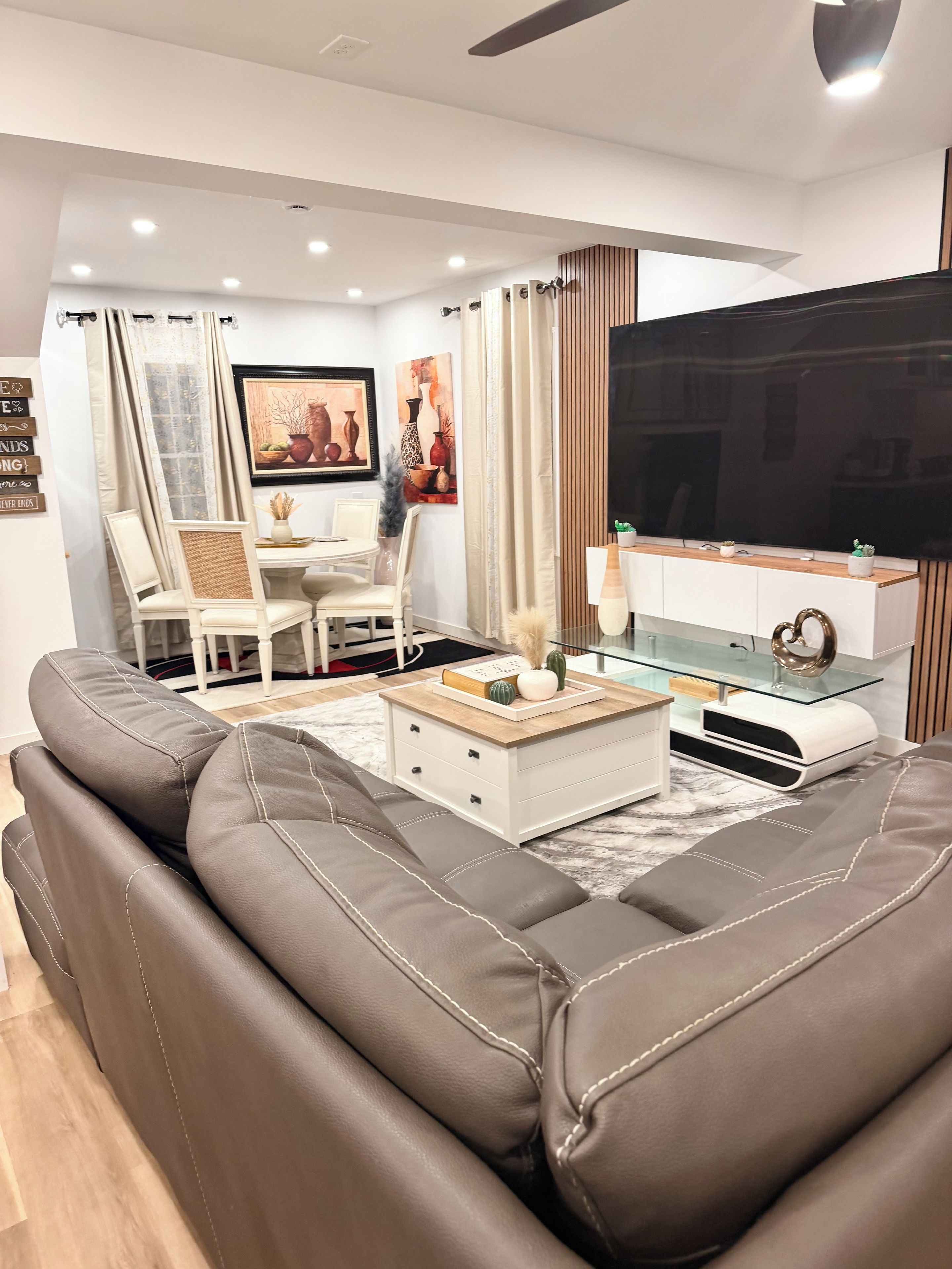A modern living room with a large sofa, a glass coffee table, a flat-screen TV, and a dining area with chairs and a table.